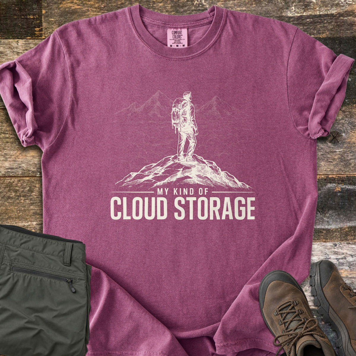 My Kind Of Cloud Storage Comfort Colors T-shirt in Berry, laid flat. A cream sketch of a backpacker on a rocky summit facing peaks above the clouds, over the words My Kind Of Cloud Storage. Folded olive hiking pants and brown hiking boots sit beside it on weathered wood.