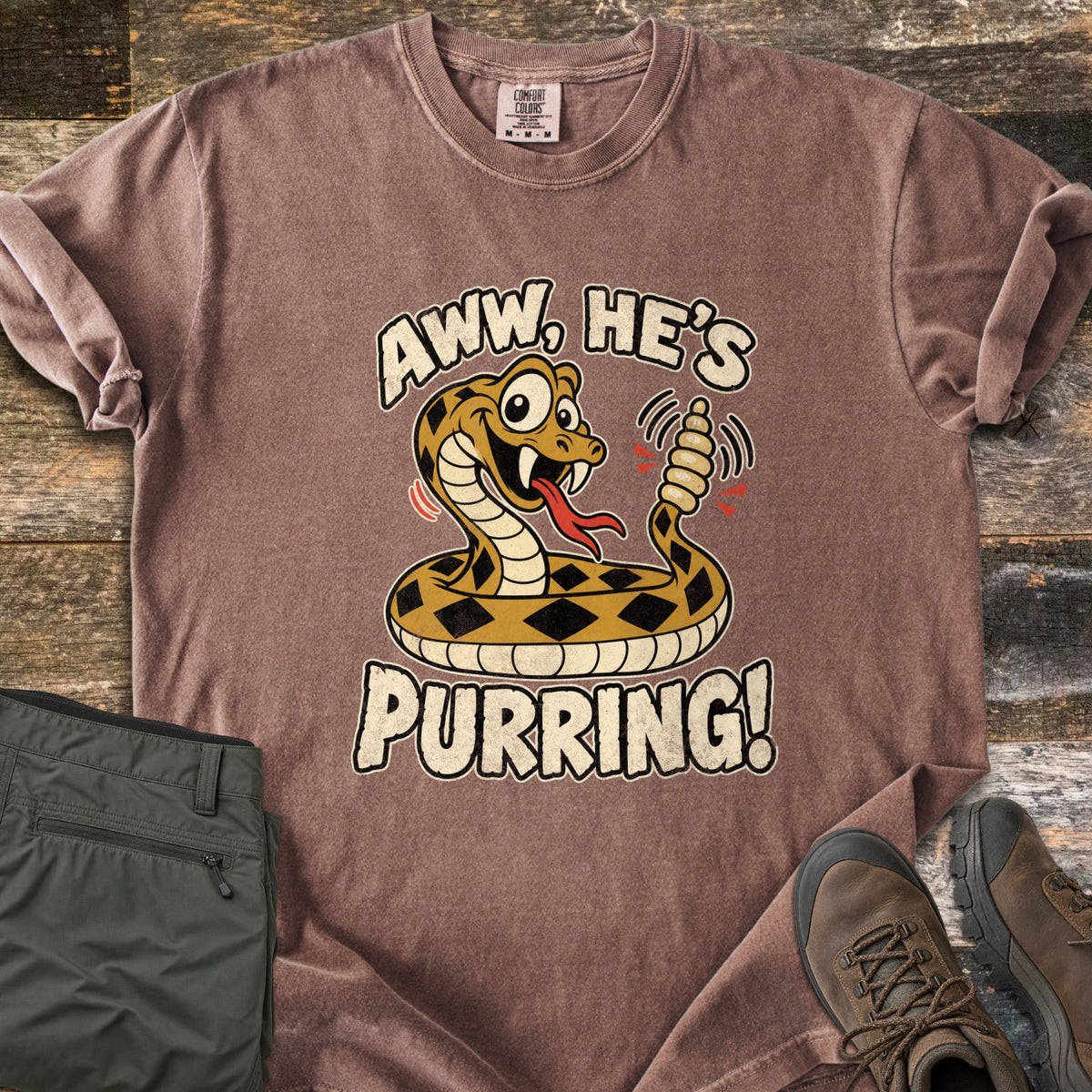 Aww He's Purring Comfort Colors T-shirt in Espresso, laid flat. A grinning, wide-eyed cartoon rattlesnake with a red forked tongue shakes its rattle between the words Aww, He's Purring! Folded olive hiking pants and brown hiking boots sit beside it on weathered wood.