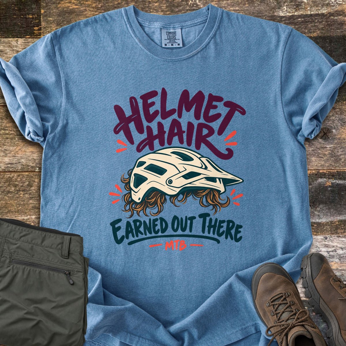 Helmet Hair Earned Out There Comfort Colors T-shirt in Blue Jean, laid flat. A cream mountain bike helmet with curly brown hair poking out below sits between the words Helmet Hair and Earned Out There, with MTB underneath. Folded olive hiking pants and brown boots lie beside it on wood planks.