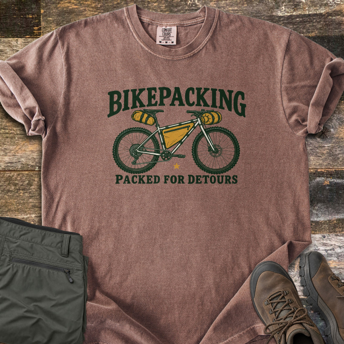 Bikepacking Packed for Detours Comfort Colors T-shirt in Espresso, laid flat. A loaded bikepacking bike with yellow bags sits between the words Bikepacking and Packed for Detours in dark green. Folded olive pants and brown hiking boots lie beside it on weathered wood planks.