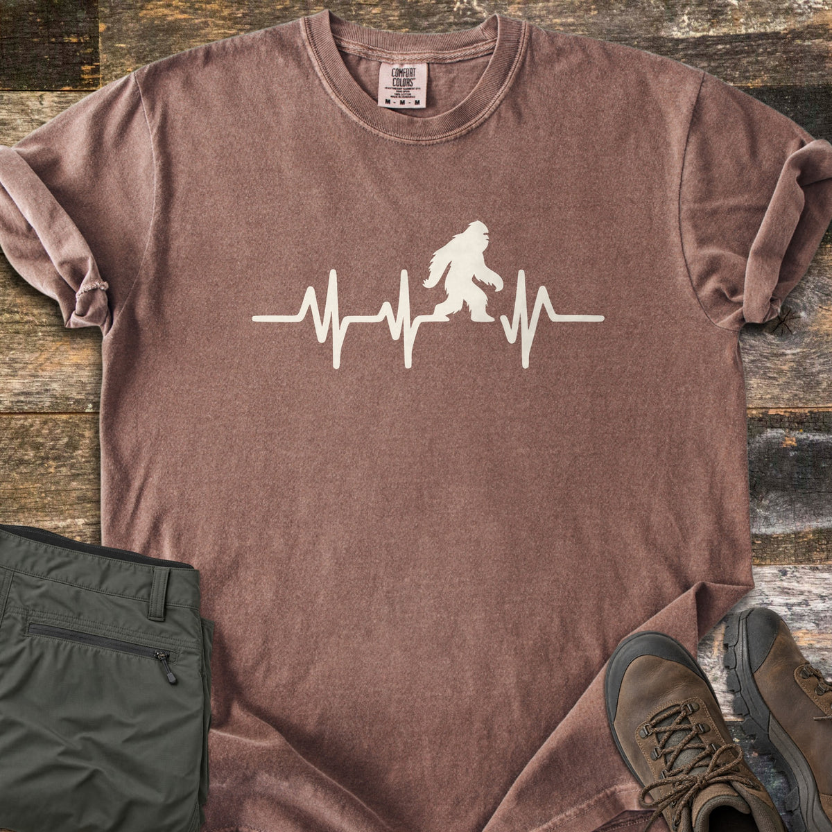 Bigfoot Heartbeat Comfort Colors T-shirt in Espresso, laid flat. A cream heartbeat line runs across the chest with a walking Bigfoot silhouette in the middle; there are no words. The shirt lies on weathered wood beside folded olive hiking pants and brown hiking boots.