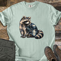 Raccoon Night Creek Comfort Colors T-shirt in Bay, laid flat. A realistic raccoon sits looking over its shoulder, its body filled with a moonlit forest creek scene with pine trees and rocks. No words. Sleeves are cuffed, on rustic wood with olive pants and brown hiking boots.