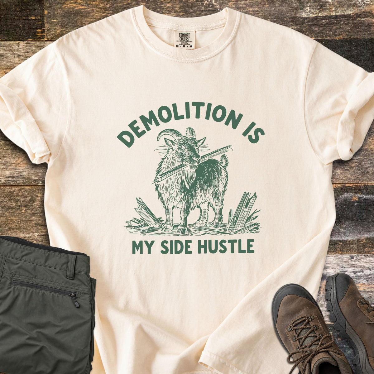 Demolition Is My Side Hustle Comfort Colors T-shirt in Ivory, laid flat. Green print of a shaggy goat holding a broken board in its mouth among splintered wood, with the words Demolition Is arched above and My Side Hustle below. Olive pants and brown hiking boots lie on weathered wood.