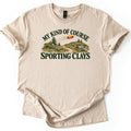 My Kind of Course Comfort Colors T-shirt in Sand, laid flat. Arched dark green text My Kind of Course tops an orange clay target flying over grassy hills with pines, rocks and a dirt path, above the words Sporting Clays. Sleeves are rolled on a white background.