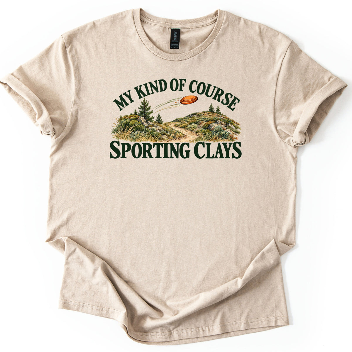 My Kind of Course Comfort Colors T-shirt in Sand, laid flat. Arched dark green text My Kind of Course tops an orange clay target flying over grassy hills with pines, rocks and a dirt path, above the words Sporting Clays. Sleeves are rolled on a white background.