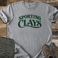 Sporting Clays Comfort Colors T-shirt in Grey, laid flat. Dark green retro lettering reads Sporting Clays, with a swash curving under the word Clays. Sleeves are rolled on a rustic wood floor beside folded olive hiking pants and brown leather hiking boots.