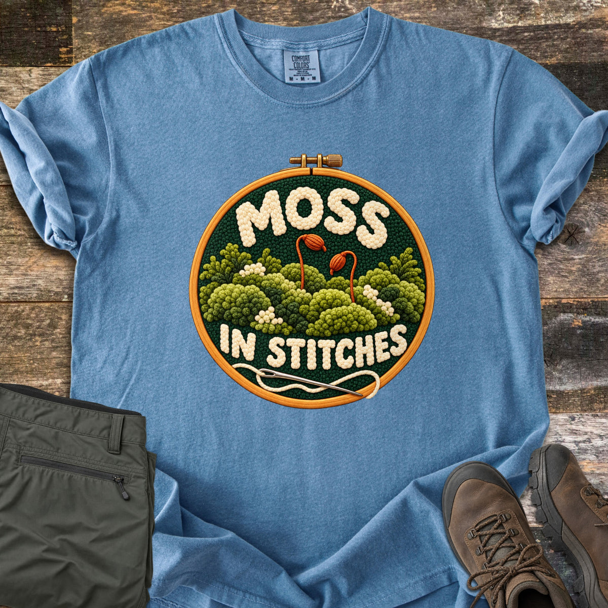 Moss in Stitches Comfort Colors T-shirt in Blue Jean, laid flat. An embroidery hoop holds stitched green moss with two orange spore capsules, the word Moss above and In Stitches below, plus a needle and thread. Folded olive pants and brown hiking boots lie beside it on weathered wood.