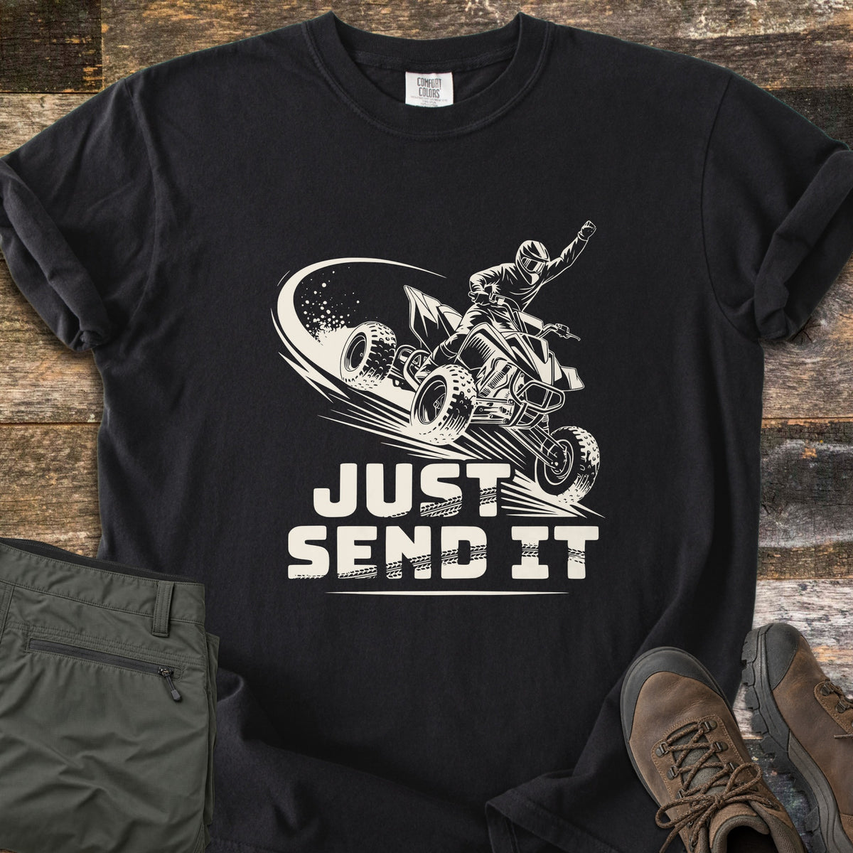 Just Send It Comfort Colors T-shirt in Black, laid flat. A cream illustration of a rider in a helmet on an airborne ATV, one fist raised, above the words Just Send It. Shown on rustic wood with olive cargo pants and brown hiking boots.