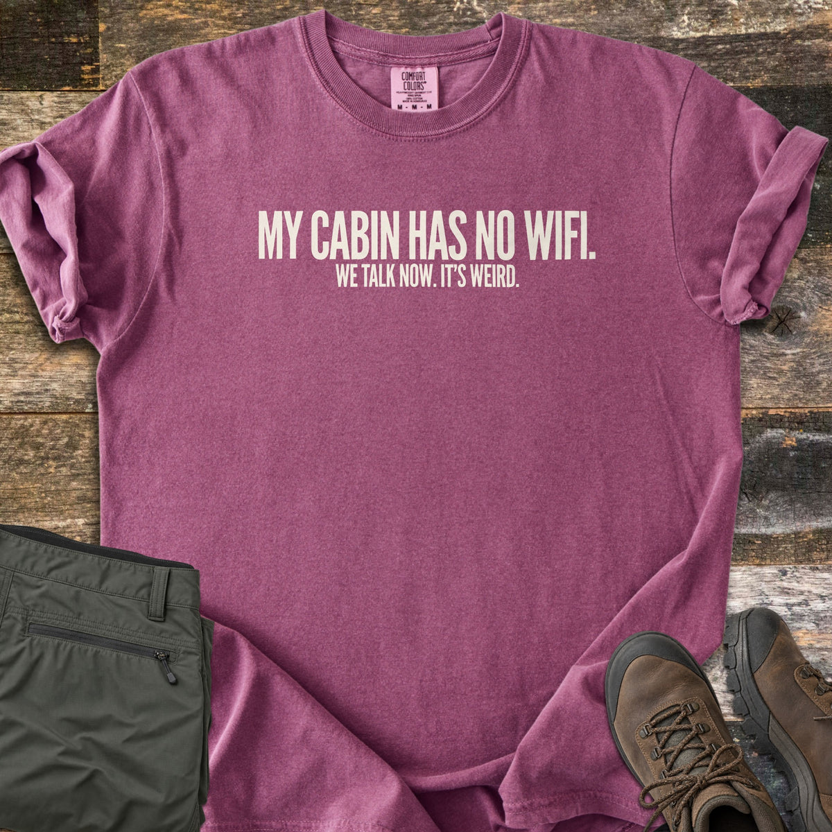My Cabin Has No Wifi Comfort Colors T-shirt in Berry, laid flat. Off-white block letters read My Cabin Has No Wifi. We Talk Now. It's Weird. The shirt has rolled sleeves and lies on weathered wood planks beside folded olive hiking pants and brown hiking boots.