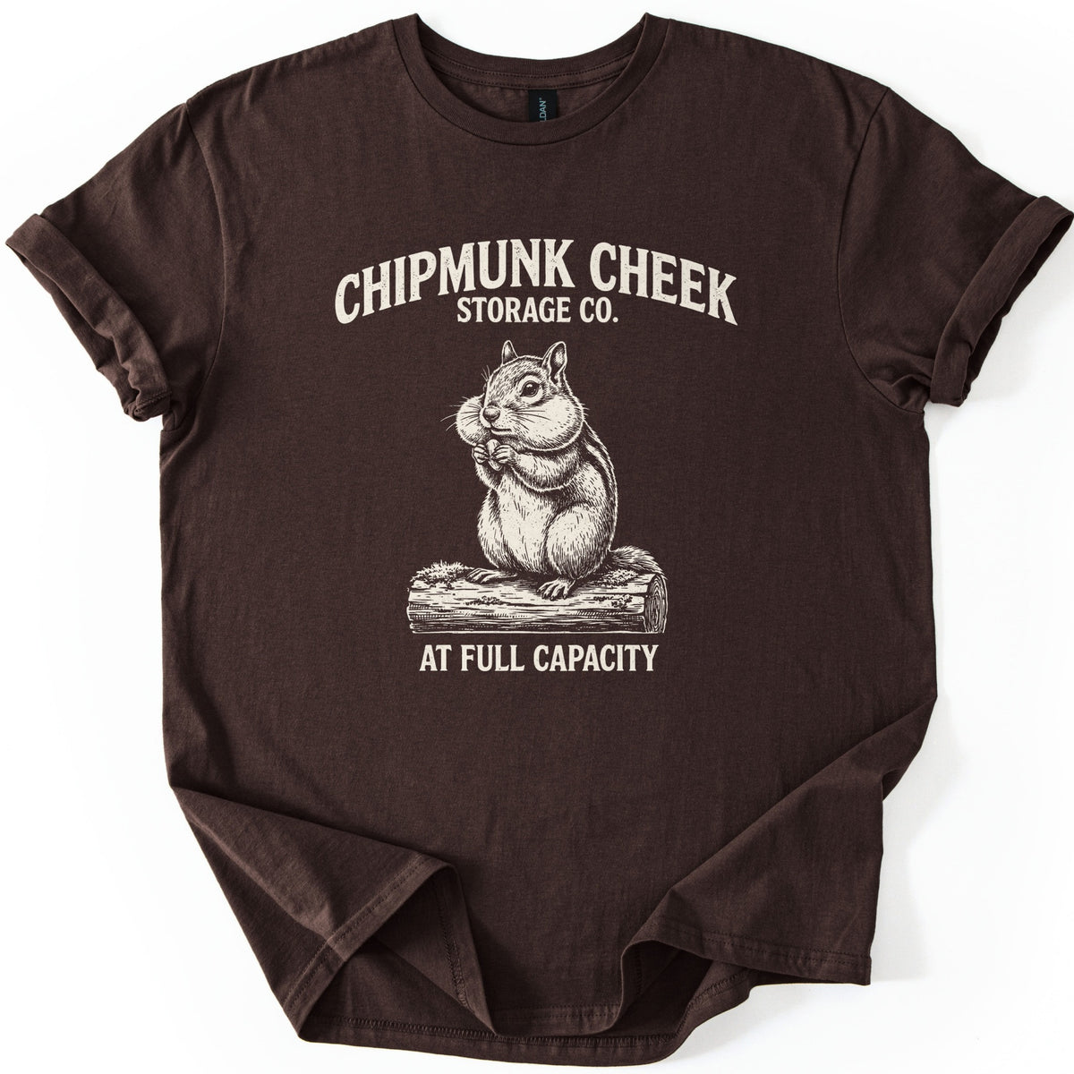 Chipmunk Cheek Storage Co Comfort Colors T-shirt in Dark Chocolate, laid flat. A cream engraving-style chipmunk with stuffed cheeks, holding a nut on a log, sits below the words Chipmunk Cheek Storage Co. and above At Full Capacity. The sleeves are rolled on a white surface.