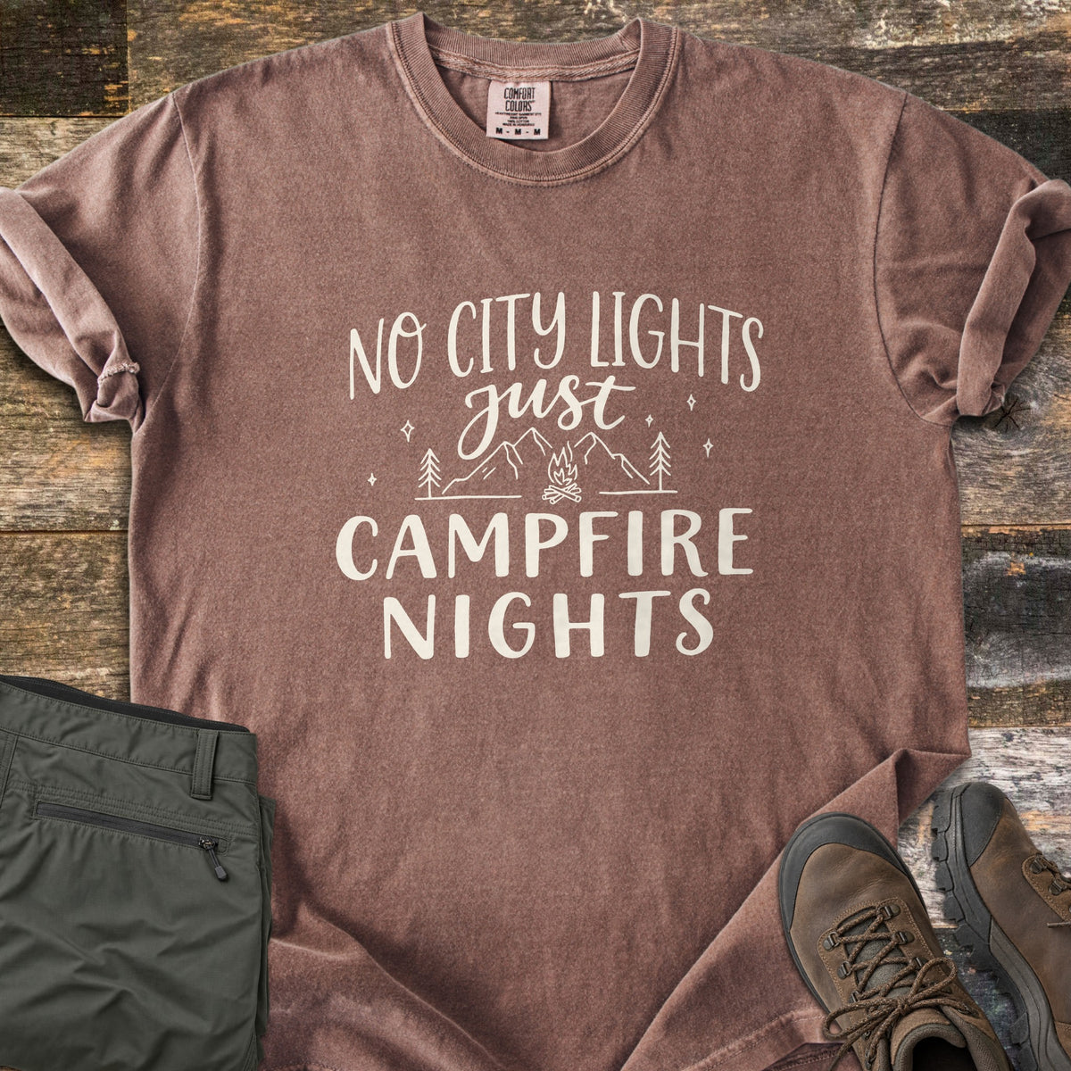Campfire Nights Comfort Colors T-shirt in Espresso, laid flat. Cream text reads No City Lights just Campfire Nights around line art of mountains, pine trees, stars and a campfire. On rustic wood planks beside folded olive hiking pants and brown hiking boots.
