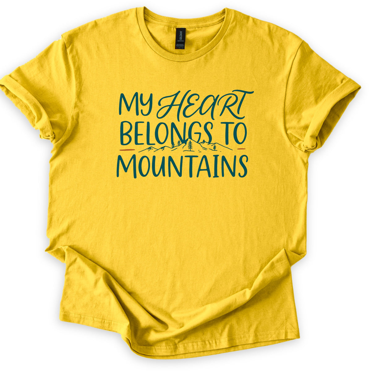 My Heart Belongs to Mountains Comfort Colors T-shirt in Daisy, laid flat. Teal lettering reads My Heart Belongs to Mountains, with Heart in script above a small line drawing of mountains and pine trees between orange dashes. The shirt has cuffed sleeves and lies on a white background.