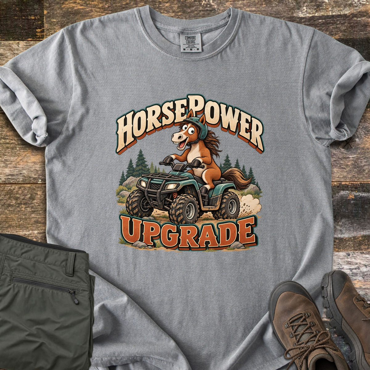 Horsepower Upgrade Comfort Colors T-shirt in Grey, laid flat. A cartoon horse in a green helmet rides a muddy green ATV past pine trees, between the words HorsePower and UPGRADE. Olive cargo pants and brown hiking boots lie beside it on weathered wood.