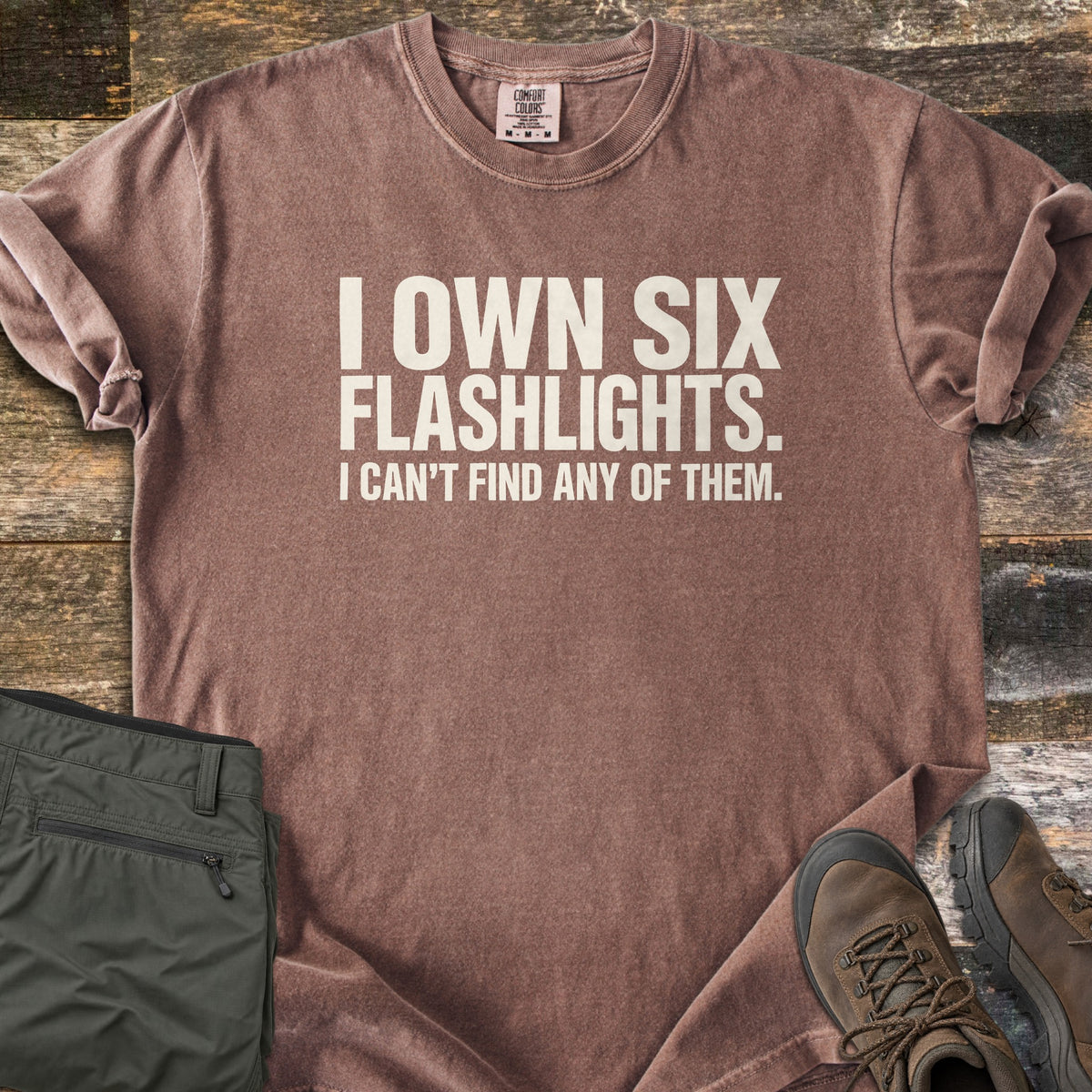 Six Flashlights Comfort Colors T-shirt in Espresso, laid flat. Bold off-white text reads I OWN SIX FLASHLIGHTS. I CAN'T FIND ANY OF THEM. The shirt has rolled sleeves and lies on weathered wood beside folded olive cargo pants and brown hiking boots.