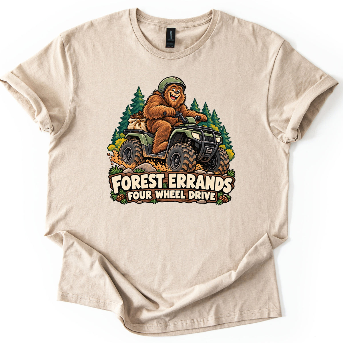 Forest Errands Four Wheel Drive Comfort Colors T-shirt in Sand, laid flat. A smiling Bigfoot wearing a green helmet drives a muddy green ATV in front of pine trees, a sack of pinecones behind him, over the words Forest Errands Four Wheel Drive. The shirt lies on a plain white surface.