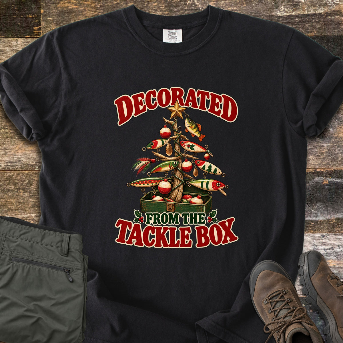Decorated From The Tackle Box Comfort Colors T-shirt in Black, laid flat. A Christmas tree made of driftwood is hung with fishing lures, bobbers and a star, rising from a green tackle box, between the words Decorated From The Tackle Box. Olive pants and brown boots sit on wood planks.
