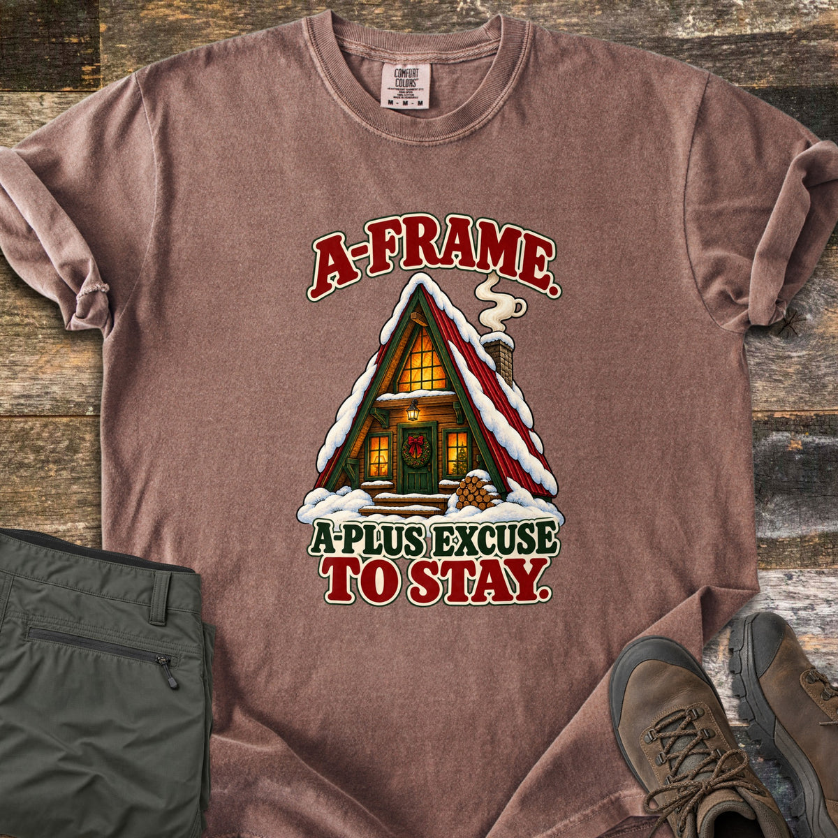 A-Frame A-Plus Excuse to Stay Comfort Colors T-shirt in Espresso, laid flat. A snowy red-roofed A-frame cabin with glowing windows, a wreathed green door, smoking chimney and stacked firewood, between the words A-Frame. A-Plus Excuse to Stay. Olive pants and brown boots lie on weathered wood.