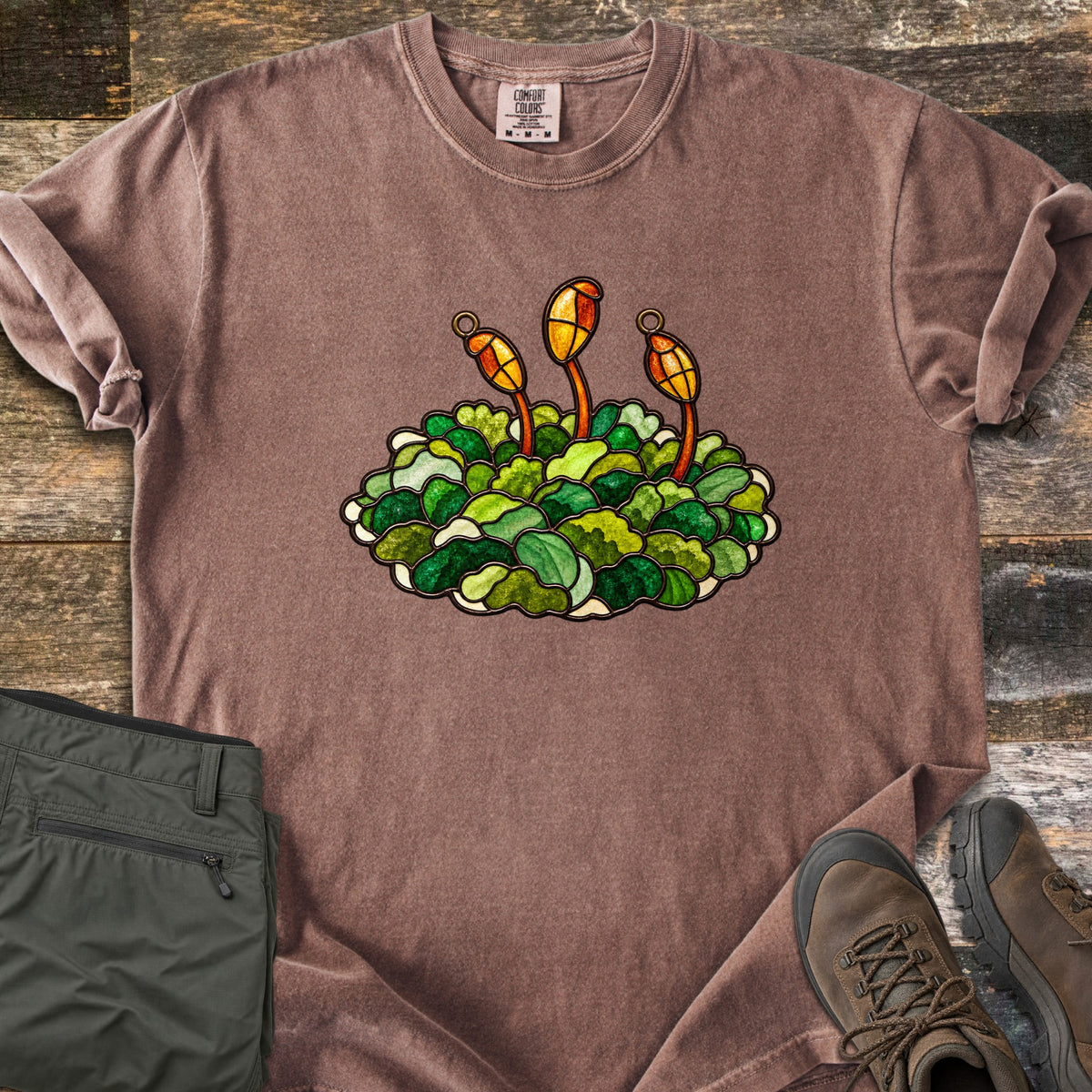 Stained Glass Moss Comfort Colors T-shirt in Espresso, laid flat. A stained glass style clump of green moss in many shades, with three amber spore capsules on thin stalks rising from it, no text. Olive cargo pants and brown hiking boots lie nearby on rustic wood.