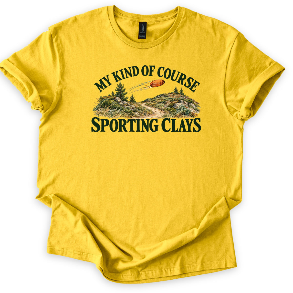 My Kind of Course Comfort Colors T-shirt in Daisy, laid flat. An orange clay target streaks over grassy hills with pine trees, rocks and a winding dirt path, framed by dark green text My Kind of Course and Sporting Clays. Sleeves are rolled on a white background.