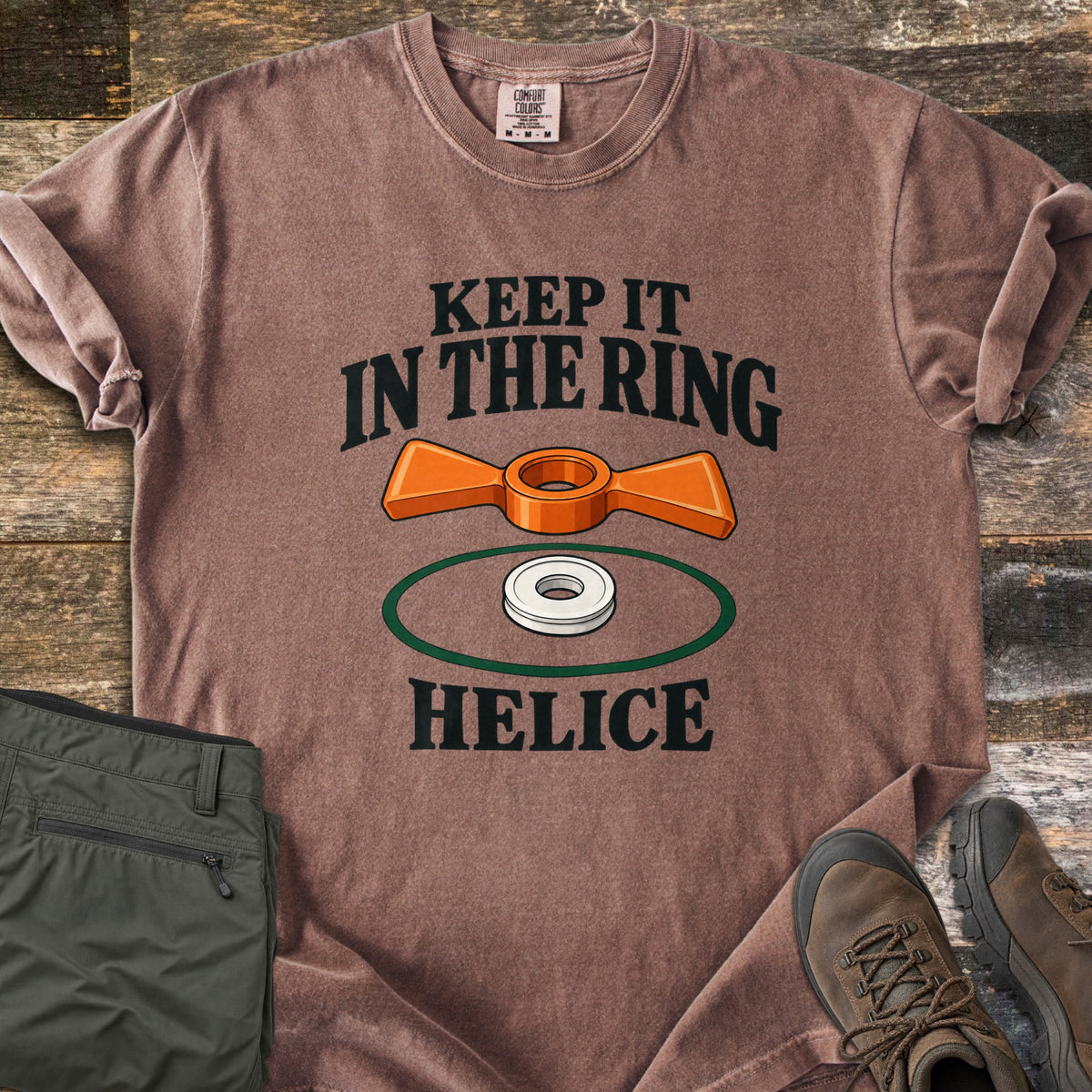 Keep It in the Ring Comfort Colors T-shirt in Espresso, laid flat. An orange winged helice target floats above a white ring inside a green circle, between the words Keep It in the Ring and Helice. Olive pants and brown hiking boots sit beside it on weathered wood.