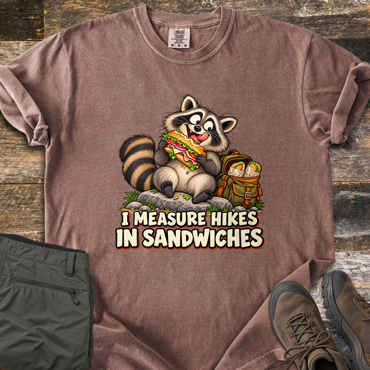 I Measure Hikes In Sandwiches Comfort Colors T-shirt in Espresso, laid flat. A cartoon raccoon sits on a rock biting a big sub sandwich beside a green backpack holding wrapped burritos, above the words I Measure Hikes In Sandwiches. Hiking pants and boots lie on a wood floor beside it.