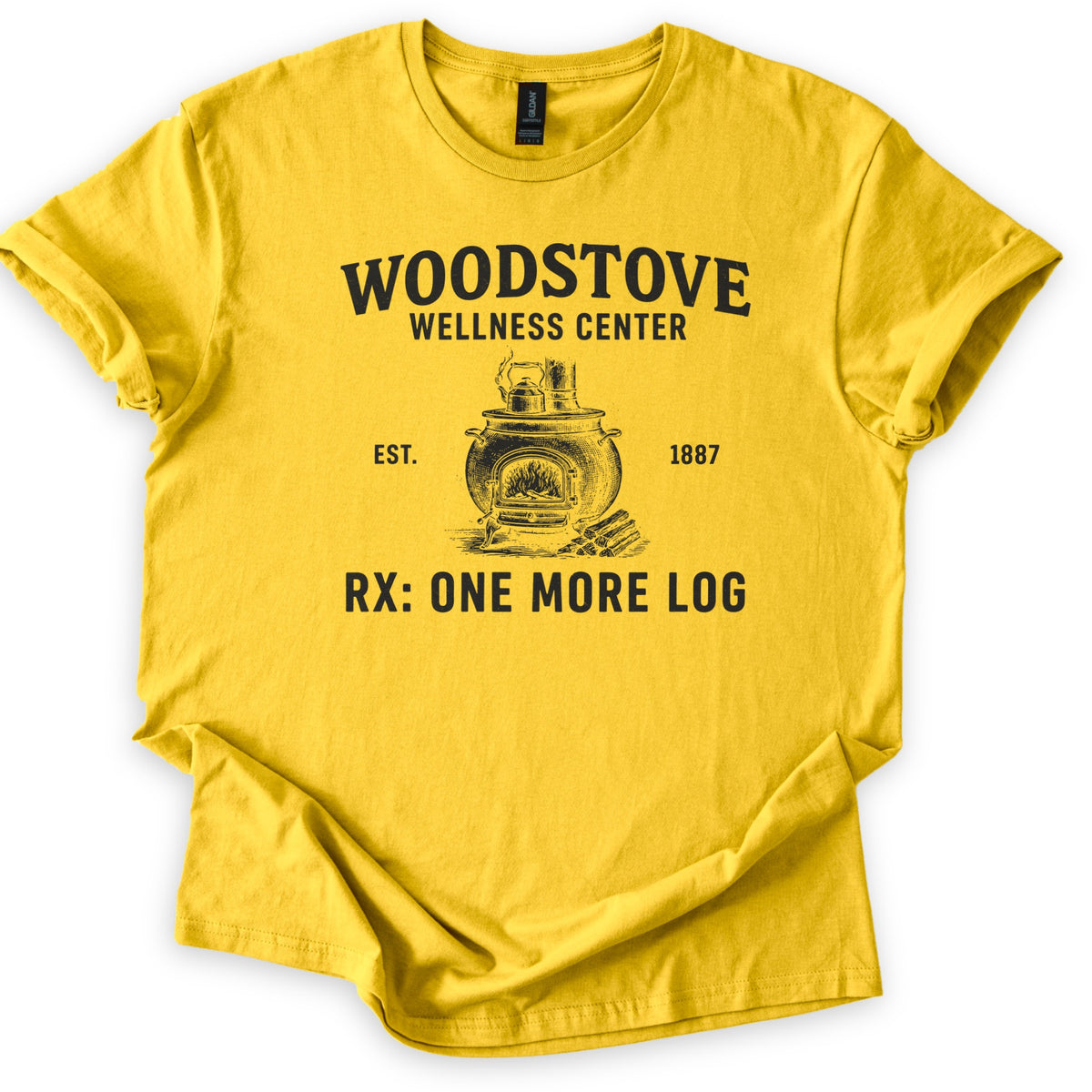Woodstove Wellness Center Comfort Colors T-shirt in Daisy, laid flat. A black sketch of a round woodstove with a kettle on top, flames in its open door and stacked logs beside it, under Woodstove Wellness Center, Est. 1887, and Rx: One More Log. Sleeves rolled, on white.
