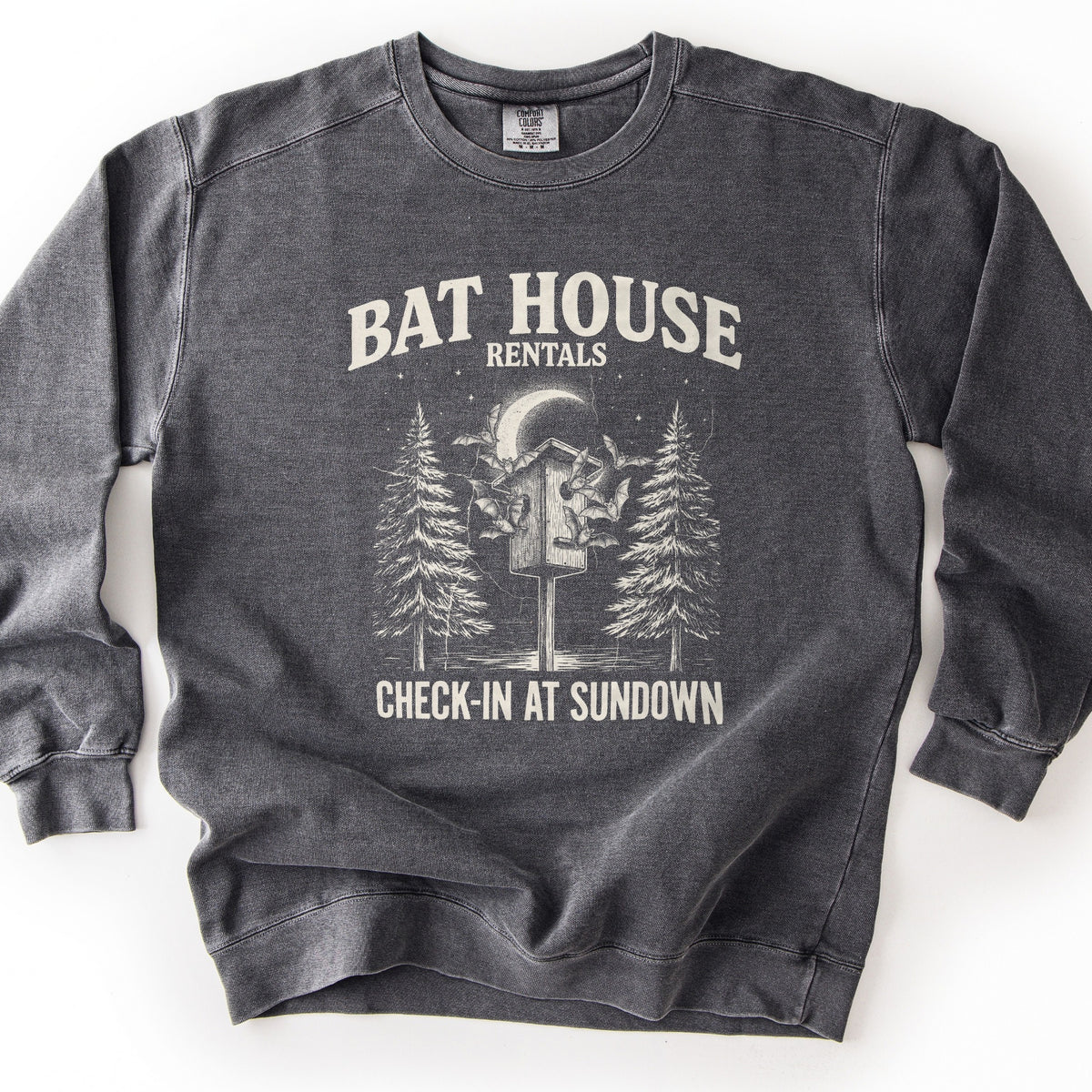 Bat House Rentals Comfort Colors crewneck sweatshirt in Pepper, laid flat. Cream print of a bat house on a post with bats flying around it under a crescent moon, between two pine trees, with the words Bat House Rentals above and Check-In At Sundown below. Shown on a white background.