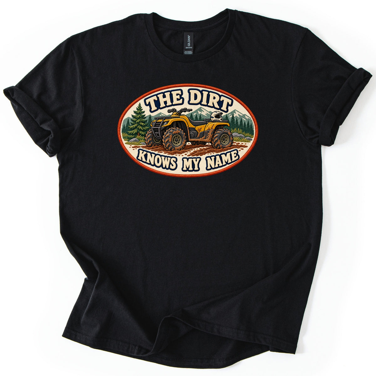 The Dirt Knows My Name Comfort Colors T-shirt in Black, laid flat. An oval patch with an orange border shows a muddy yellow ATV on a dirt trail by pine trees and mountains, with THE DIRT above and KNOWS MY NAME below. The shirt lies on a white surface with sleeves rolled.
