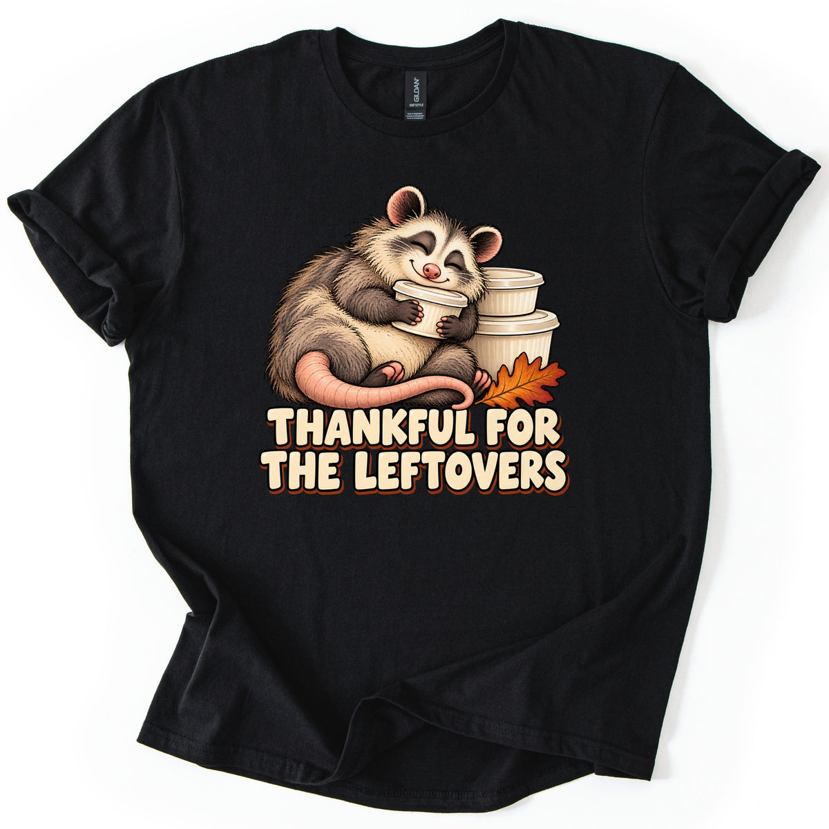 Thankful For The Leftovers Comfort Colors T-shirt in Black, laid flat. A content opossum with a pink tail hugs a white leftovers container next to stacked containers and an orange autumn leaf, above cream letters reading Thankful For The Leftovers. Sleeves cuffed on white.