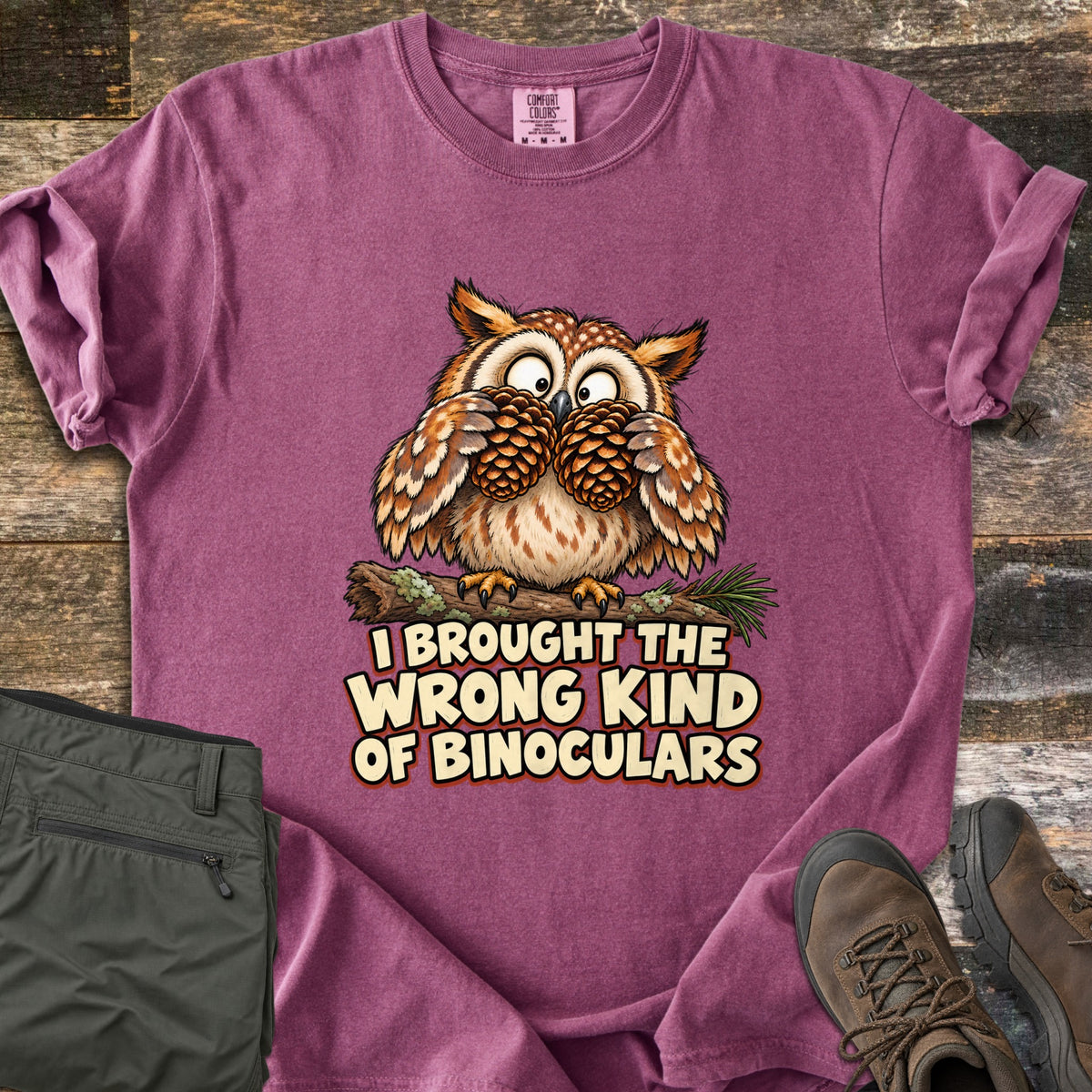 I Brought The Wrong Kind Of Binoculars Comfort Colors T-shirt in Berry, laid flat. A wide-eyed cartoon owl on a mossy branch holds two pinecones to its eyes like binoculars above the words I Brought The Wrong Kind Of Binoculars. Olive hiking pants and brown hiking boots lie beside it on weathered wood.