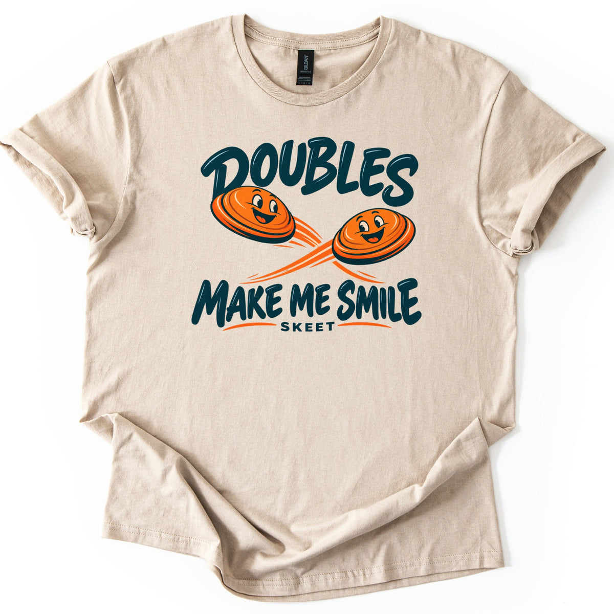 Doubles Make Me Smile Comfort Colors T-shirt in Sand, laid flat. Two smiling orange clay targets with motion streaks fly between the words Doubles and Make Me Smile in dark teal, with Skeet in small letters below. The shirt has rolled sleeves on a white background.