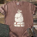 Backpack And Bedroll Comfort Colors T-shirt in Espresso, laid flat. A cream canvas backpack hanging from a wall peg above a bedroll tied with rust-orange straps. Olive hiking pants and brown hiking boots sit beside it on weathered wood planks.