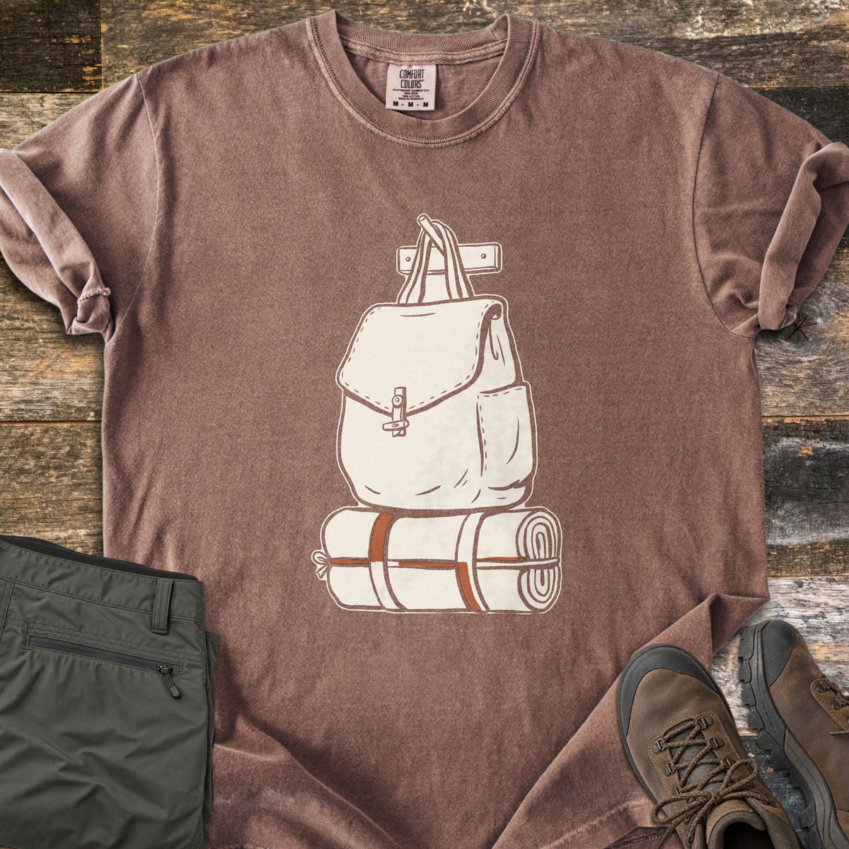 Backpack And Bedroll Comfort Colors T-shirt in Espresso, laid flat. A cream canvas backpack hanging from a wall peg above a bedroll tied with rust-orange straps. Olive hiking pants and brown hiking boots sit beside it on weathered wood planks.