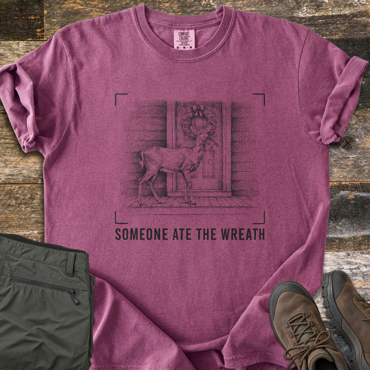 Someone Ate The Wreath Comfort Colors T-shirt in Berry, laid flat. A sketched deer nibbles a wreath on a cabin's front door, framed by corner marks above the words Someone Ate The Wreath. Shown on rustic wood planks with folded olive pants and brown hiking boots.