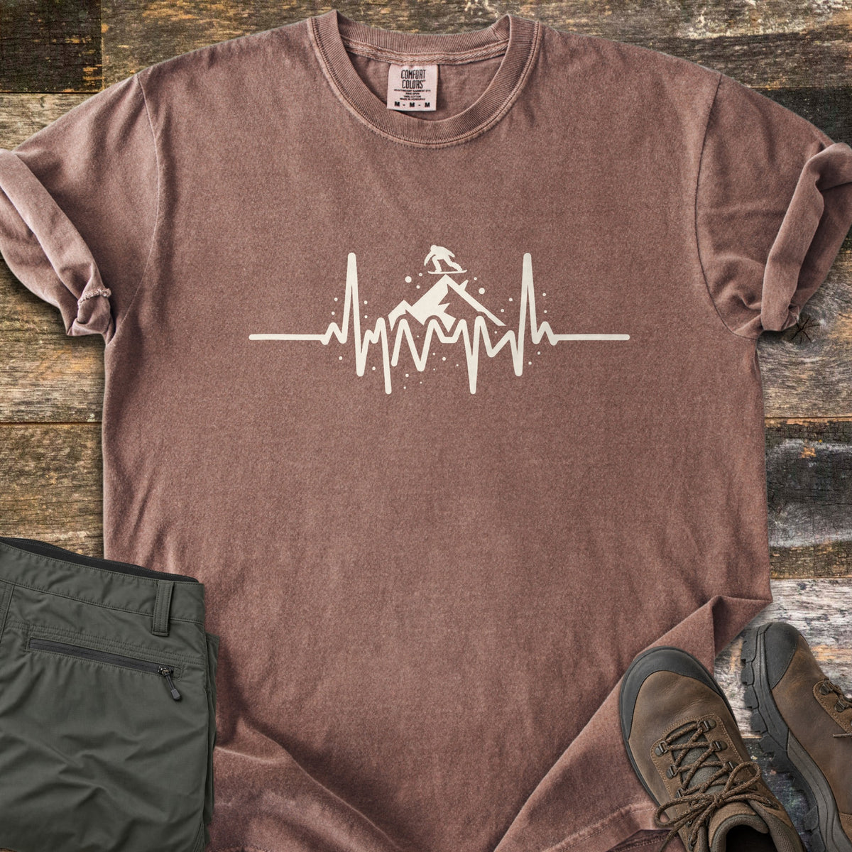 Snow Boarder Heartbeat Comfort Colors T-shirt in Espresso, laid flat. A cream heartbeat line rises into a mountain peak with a snowboarder riding on top, surrounded by snow dots. Folded olive hiking pants and brown leather boots sit beside it on weathered wood planks.