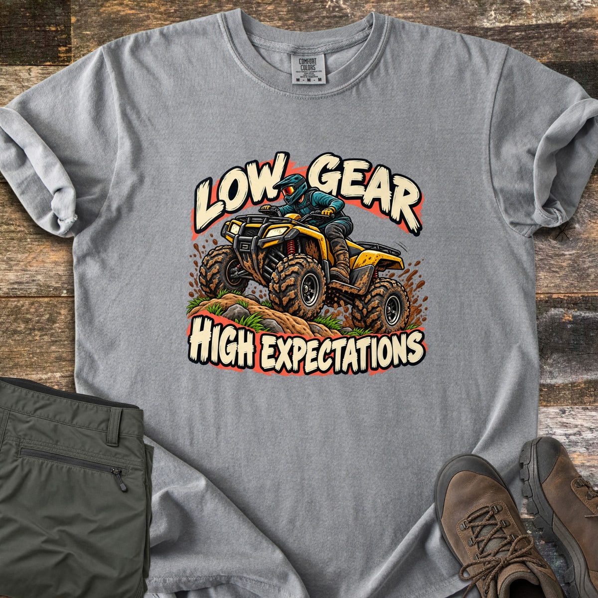 Low Gear High Expectations Comfort Colors T-shirt in Grey, laid flat. A rider in a teal helmet drives a muddy yellow ATV over a dirt mound, between the words Low Gear and High Expectations. Folded green cargo pants and brown hiking boots lie on weathered wood.