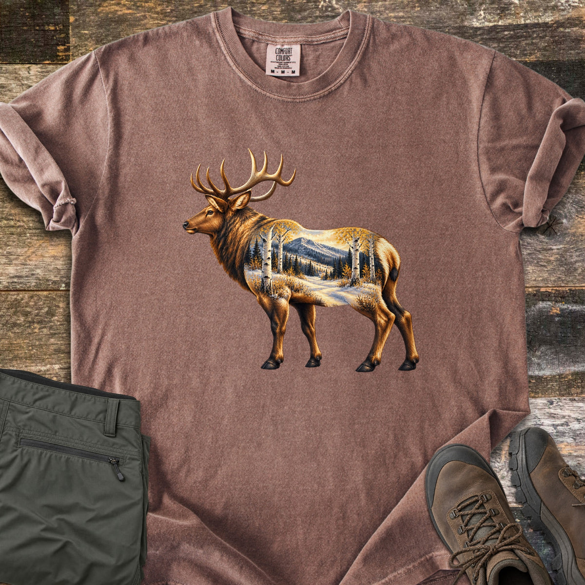 Aspen Pass Elk Comfort Colors T-shirt in Espresso, laid flat. A bull elk stands in profile, its body showing a snowy valley with white aspen trunks, pines and a mountain. Laid on rustic wood planks beside folded olive hiking pants and brown leather hiking boots.