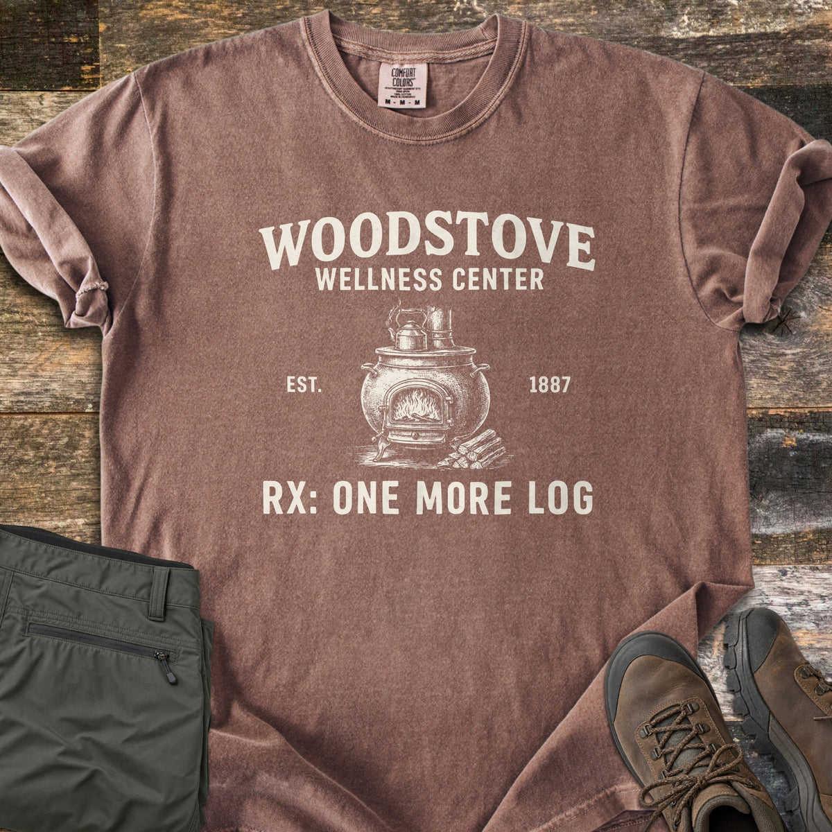Woodstove Wellness Center Comfort Colors T-shirt in Espresso, laid flat. Cream print of a potbelly woodstove with a fire, a kettle on top and logs beside it, under Woodstove Wellness Center, flanked by Est. 1887, above Rx: One More Log. On wood planks with olive pants and brown hiking boots.