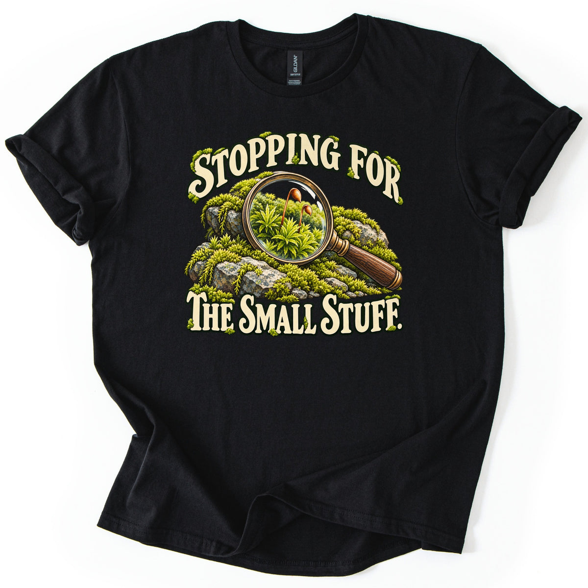 Stopping for the Small Stuff Comfort Colors T-shirt in Black, laid flat. Cream lettering reads Stopping For The Small Stuff. around a magnifying glass on mossy rocks, its lens showing green moss and tiny spore capsules. Sleeves are rolled on a white background.