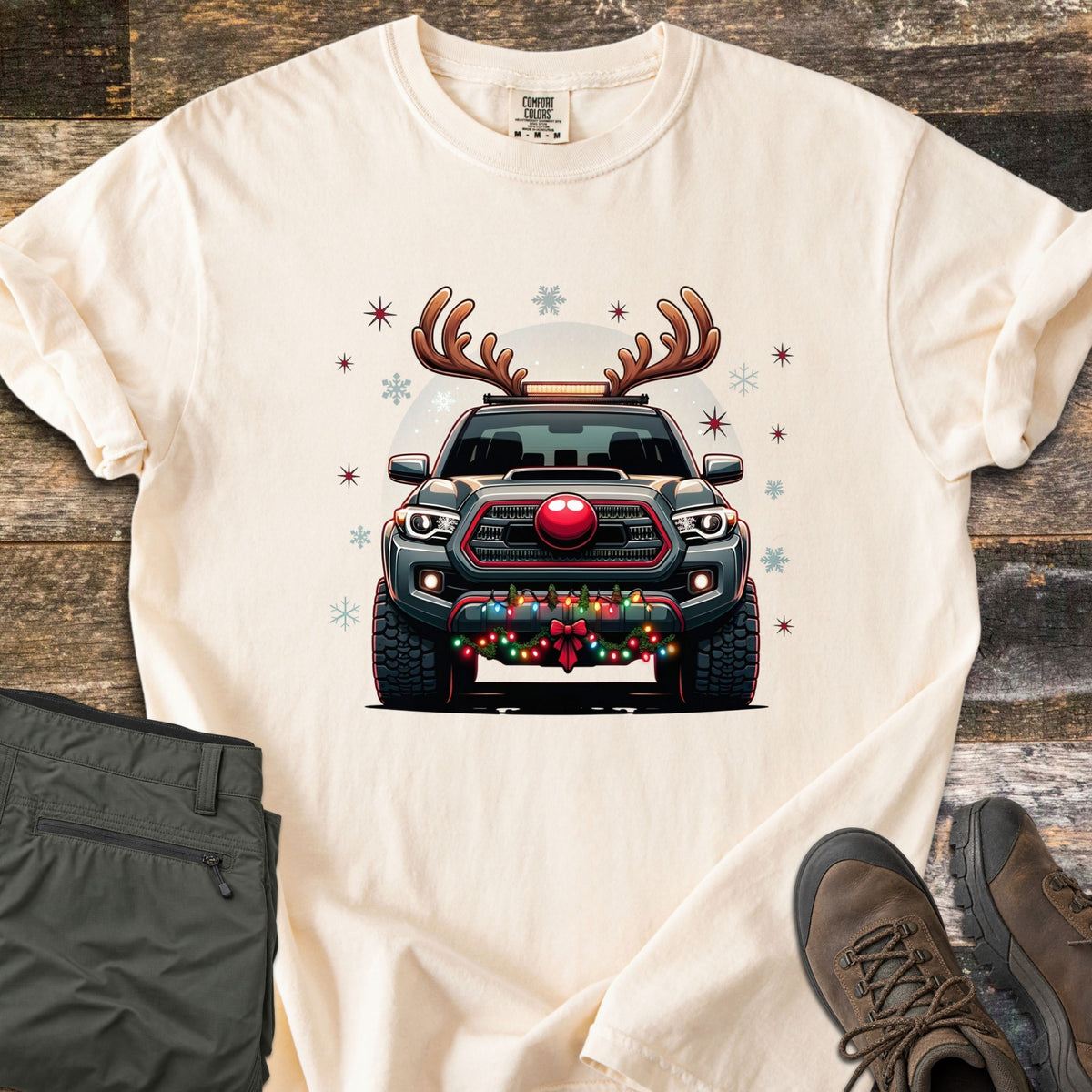 Santa Truck Comfort Colors T-shirt in Ivory, laid flat. A gray pickup truck wearing reindeer antlers and a red nose, with string lights and a red bow on the bumper, among stars and snowflakes. Laid on a wood floor with olive pants and brown hiking boots.