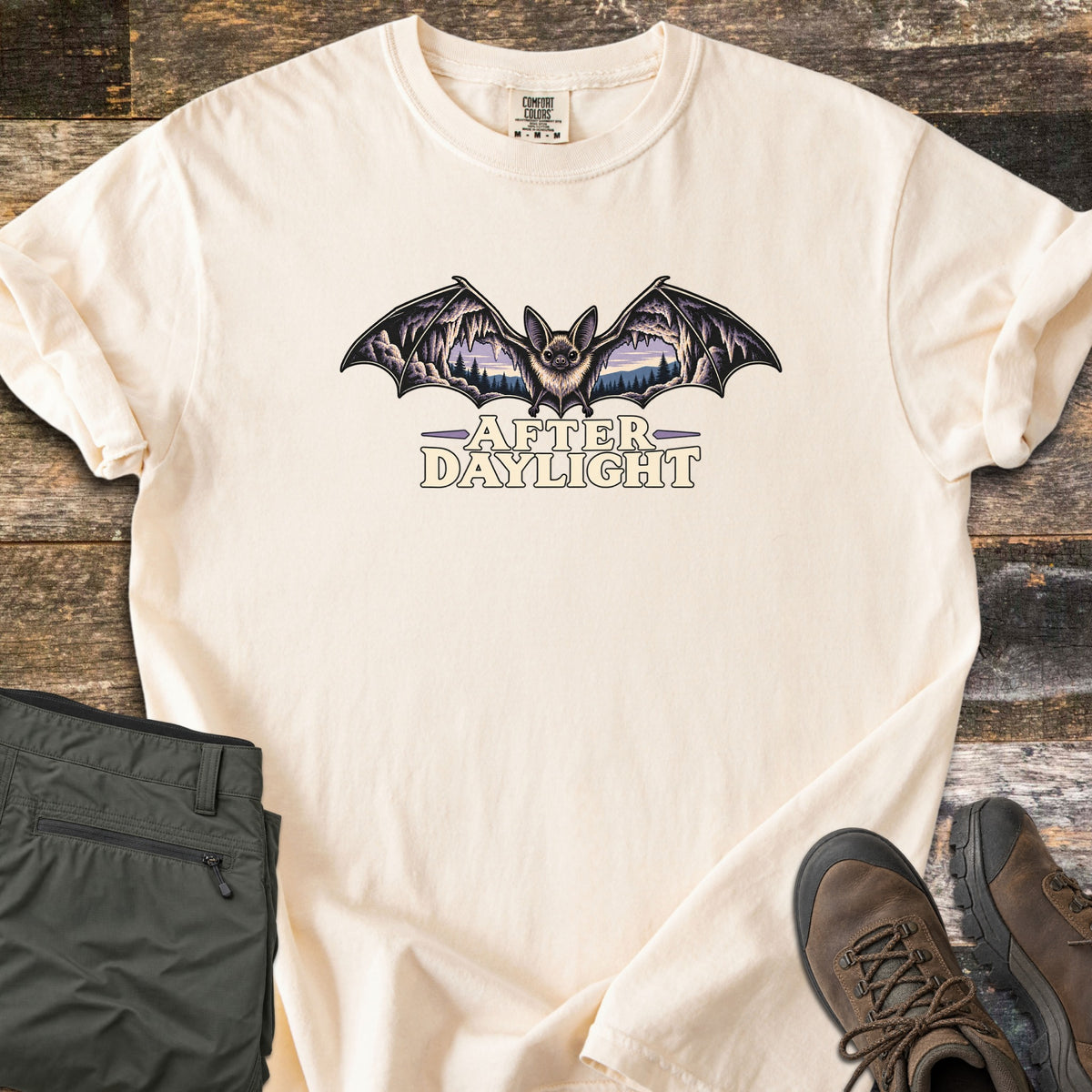 After Daylight Comfort Colors T-shirt in Ivory, laid flat. A bat spreads its wings, each framing a cave view of stalactites and pine forest at dusk, above the words After Daylight. Olive hiking pants and brown hiking boots lie beside it on dark wood planks.