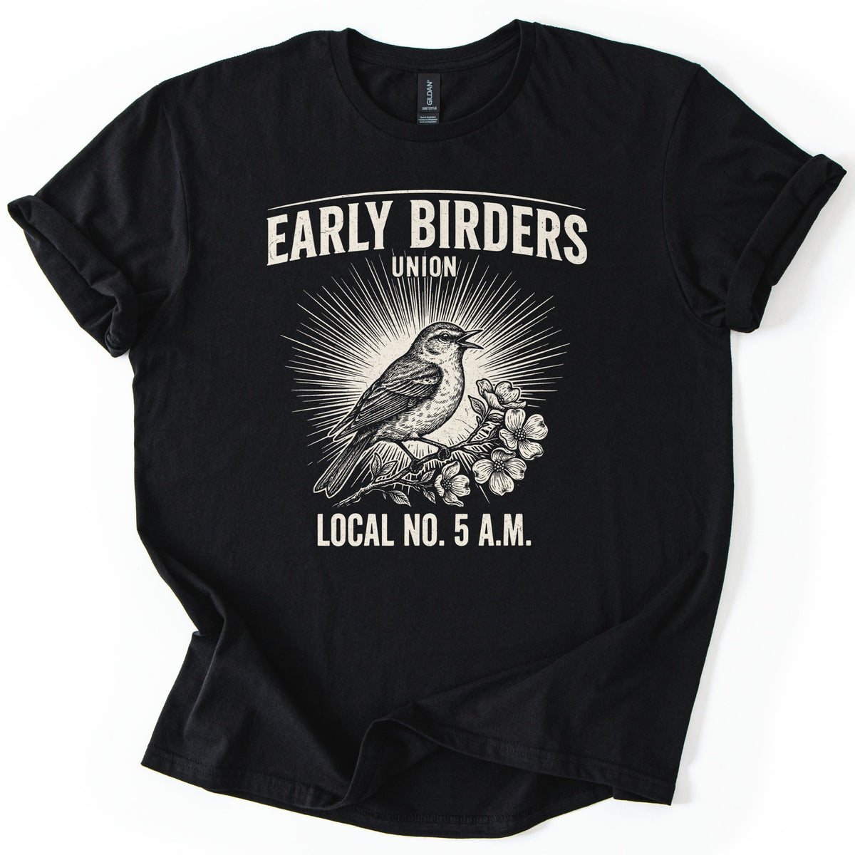 Early Birders Union Comfort Colors T-shirt in Black, laid flat. A cream engraving-style songbird perched on a blossoming branch sings in front of sunburst rays, under the words Early Birders Union and above Local No. 5 A.M. The shirt lies on a white surface with its sleeves cuffed.