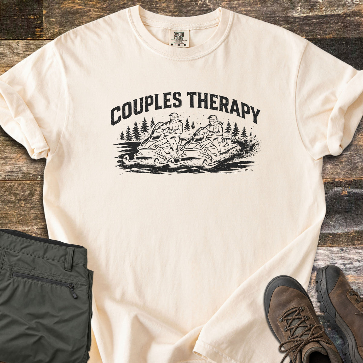 Couples Therapy Snowmobile Comfort Colors T-shirt in Ivory, laid flat. The words COUPLES THERAPY arch in black over two riders on snowmobiles kicking up snow among pine trees. Olive cargo pants and brown hiking boots lie beside it on dark weathered wood.
