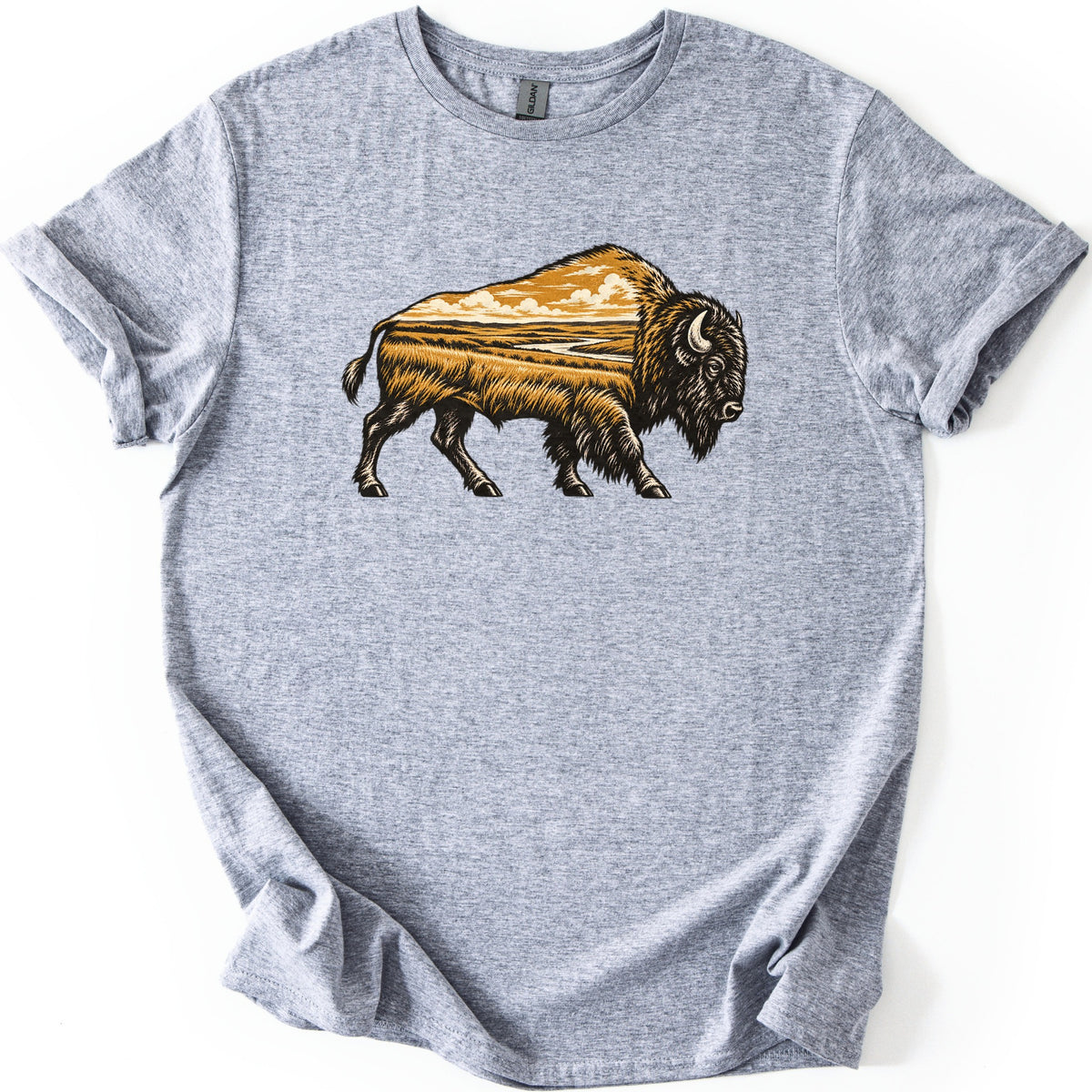 Prairie Bison Comfort Colors T-shirt in Sport Grey, laid flat. A woodcut-style bison walks in side profile, its body filled with a golden prairie scene of tall grass, a winding river and clouds. No words. The shirt has cuffed sleeves on a white background.