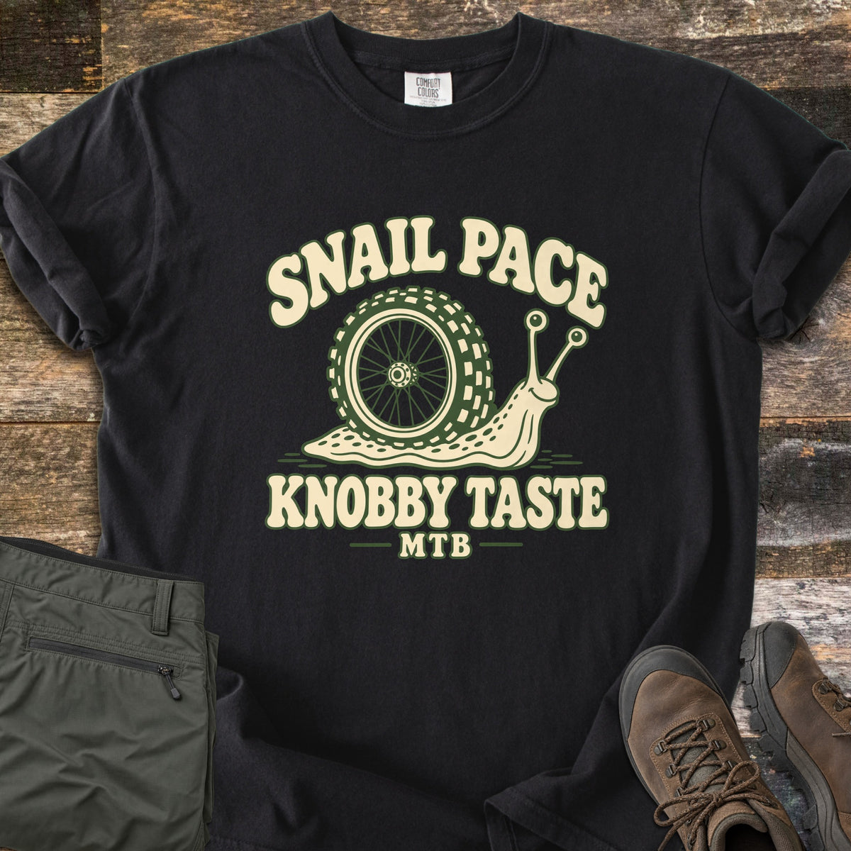 Snail Pace Knobby Taste Comfort Colors T-shirt in Black, laid flat. A cartoon snail whose shell is a knobby mountain bike tire sits between the words Snail Pace and Knobby Taste, with MTB below. It lies on weathered wood beside folded olive hiking pants and brown hiking boots.