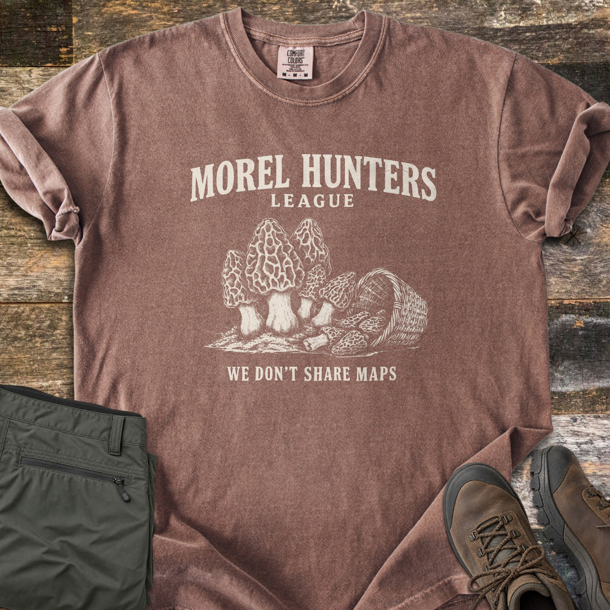 Morel Hunters League Comfort Colors T-shirt in Espresso, laid flat. Cream drawing of morel mushrooms and an overturned wicker basket of morels, between Morel Hunters League and We Don't Share Maps. Laid on rustic wood planks beside folded olive hiking pants and brown hiking boots.