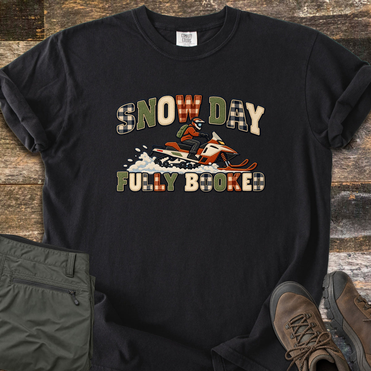 Snow Day Fully Booked Comfort Colors T-shirt in Black, laid flat. A rider in an orange jacket and helmet speeds on an orange and white snowmobile through snow between the words Snow Day and Fully Booked in green, cream and plaid letters. Olive pants and brown boots sit on a wood floor.