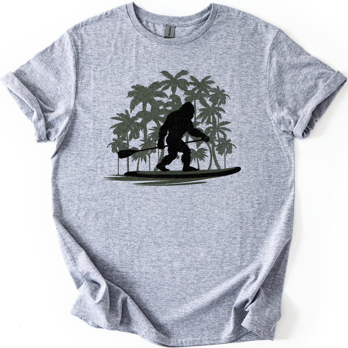 Paddle Board Squatch Comfort Colors T-shirt in Sport Grey, laid flat. A black silhouette of Bigfoot stands on a paddle board holding a paddle, in front of a cluster of olive green palm trees, with no words printed. The shirt lies on a white background with rolled sleeves.