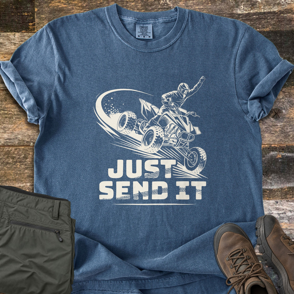Just Send It Comfort Colors T-shirt in Denim, laid flat. A cream illustration of a rider in a helmet on an airborne ATV, one fist raised, above the words Just Send It. Shown on rustic wood with olive cargo pants and brown hiking boots.