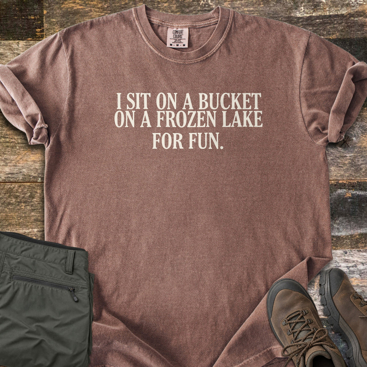 Bucket On A Frozen Lake Comfort Colors T-shirt in Espresso, laid flat. Cream serif text reads I SIT ON A BUCKET ON A FROZEN LAKE FOR FUN. The shirt has rolled sleeves and lies on weathered wood planks beside folded olive hiking pants and brown hiking boots.